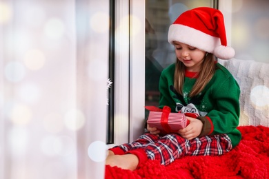Photo of Cute little girl in Santa hat holding Christmas present on window sill at home