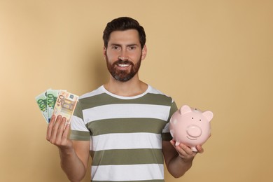 Happy man with money and ceramic piggy bank on beige background