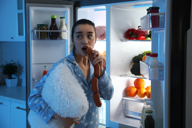 Young woman with pillow eating sausages near open refrigerator at night