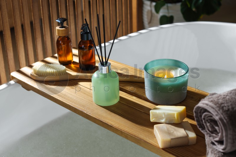 Wooden bath tray with candle, air freshener and bathroom amenities on tub indoors