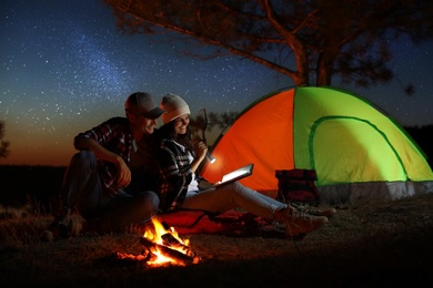 Photo of Couple with flashlight reading book near bonfire at night. Camping season