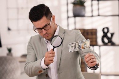Photo of Expert authenticating 100 dollar banknote with magnifying glass in office, focus on hand. Fake money concept