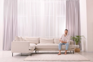Photo of Happy man drinking coffee while resting on sofa near window with beautiful curtains in living room