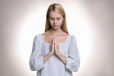 Religious young woman with clasped hands praying against light background