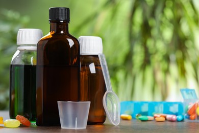 Photo of Bottles of syrup, measuring cup, dosing spoon and pills on wooden table against blurred background, space for text. Cold medicine