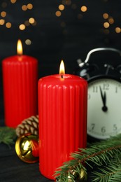 Burning candles, festive decor and alarm clock on wooden table against blurred Christmas lights