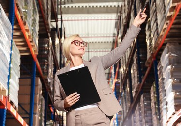 Happy manager holding clipboard in warehouse with lots of products, low angle view