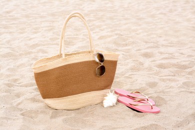 Stylish straw bag with sunglasses, flip flops and seashell on sand outdoors