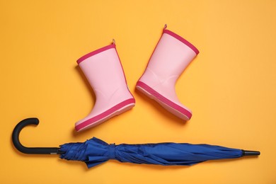 Pink rubber boots near blue umbrella on orange background, flat lay