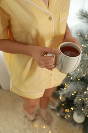 Woman with cup of tea near Christmas tree indoors, closeup