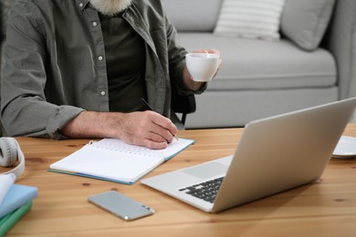 Man with laptop, notebook and cup of drink learning at wooden table indoors, closeup