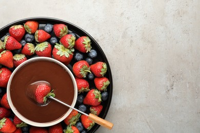 Photo of Fondue fork with strawberry in bowl of melted chocolate surrounded by different berries on light table, top view. Space for text