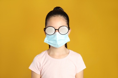 Little girl with foggy glasses caused by wearing medical face mask on yellow background. Protective measure during coronavirus pandemic