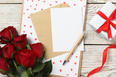 Flat lay composition with blank greeting card, gift and roses on white wooden table. Valentine's day celebration