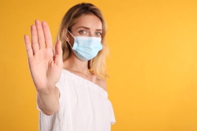 Woman in protective face mask showing stop gesture on yellow background, focus on hand. Prevent spreading of coronavirus