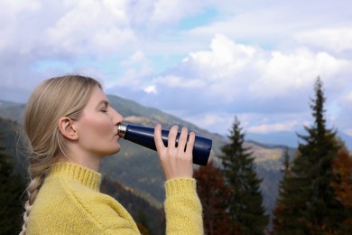 Young woman drinking hot beverage from thermo bottle in mountains