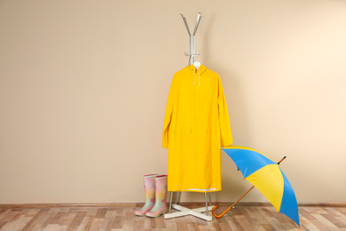 Umbrella, rain coat and rubber boots near beige wall. Space for text