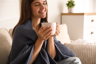 Woman covered with warm dark blue plaid enjoying hot drink on sofa at home