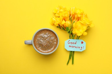 Cup of aromatic coffee, beautiful freesias and Good Morning note on yellow background, flat lay