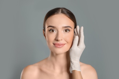 Doctor examining woman's face before plastic surgery on grey background