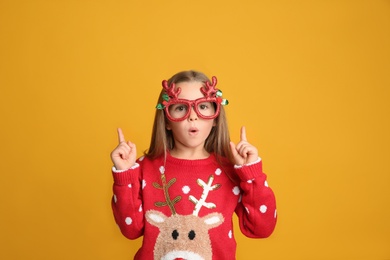 Surprised little girl in Christmas sweater and party glasses pointing on yellow background