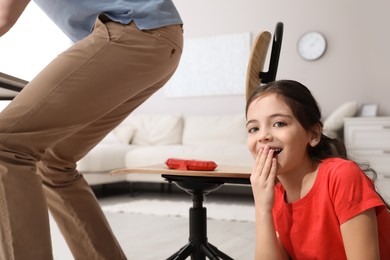 Photo of Cute little girl putting whoopee cushion on father's chair while he sitting down at home, closeup