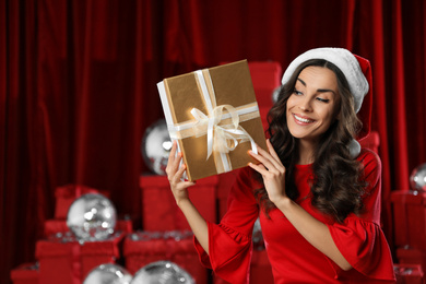 Beautiful woman in Christmas costume with gift near pile of presents indoors