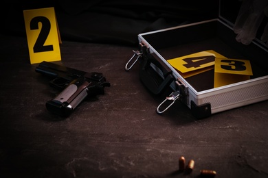 Gun and open case with evidence markers on black slate table. Crime scene