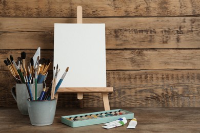 Easel with blank canvas, paints and brushes on wooden table. Space for text