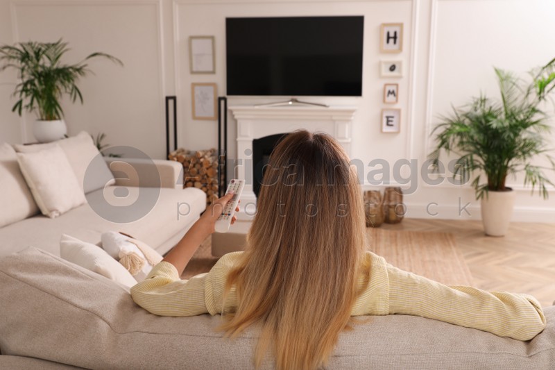 Photo of Young woman watching television at home, back view. Living room interior with TV on fireplace