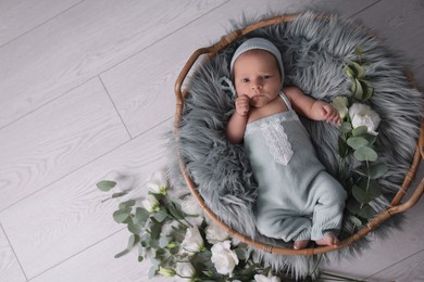 Adorable newborn baby lying in basket with faux fur and flowers on floor, top view. Space for text 