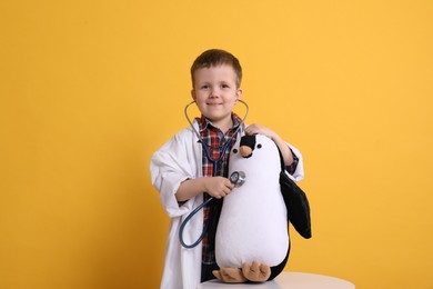 Cute little boy in pediatrician's uniform playing with stethoscope and toy penguin on yellow background