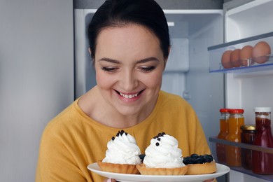 Happy overweight woman with cakes near fridge in kitchen