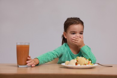 Cute little girl covering mouth and refusing to drink juice at table on grey background