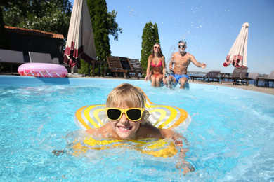 Little boy swimming with inflatable ring near his parents in outdoor pool on sunny summer day