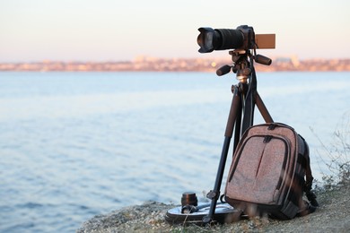 Photo of Tripod with modern camera and set of photography equipment on rocky river coast. Space for text