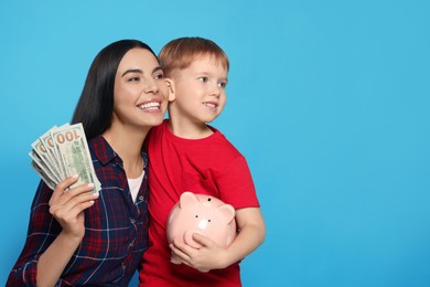 Mother holding money and her son with piggy bank on light blue background, space for text