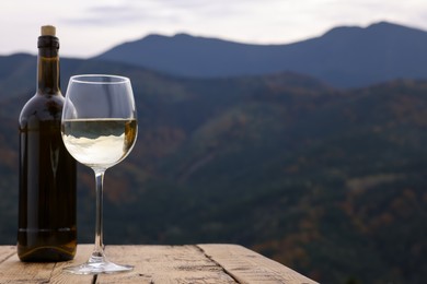 Photo of Glass and bottle of tasty wine on wooden table against mountain landscape. Space for text