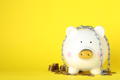 Piggy bank with steel chain and coins on yellow background, space for text. Money safety concept