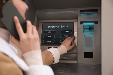 Image of Young woman talking by mobile phone while using cash machine for money withdrawal outdoors, closeup