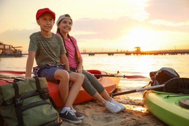 Happy children sitting on kayak near river at sunset. Summer camp