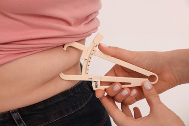 Nutritionist measuring overweight woman's body fat layer with caliper on white background, closeup