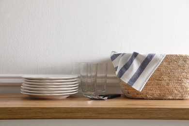 Kitchen towel in wicker basket and clean dishware on wooden table near white wall