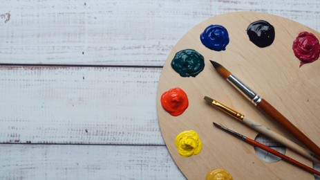 Photo of Artist's palette with samples of colorful paints and brushes on white wooden table, top view. Space for text