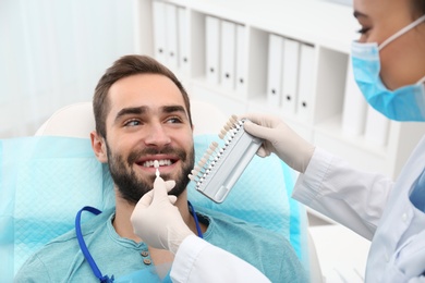 Dentist matching young man's teeth color with palette in office