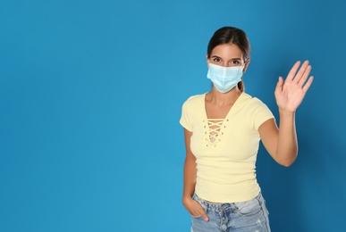 Woman in protective mask showing hello gesture on blue background, space for text. Keeping social distance during coronavirus pandemic