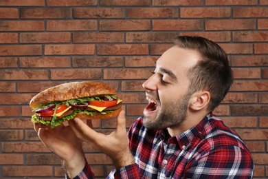 Young hungry man eating tasty sandwich on brick wall background
