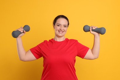 Happy overweight woman doing exercise with dumbbells on orange background