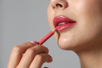 Young woman applying beautiful pink lip pencil on grey background, closeup