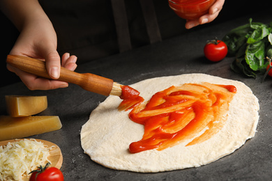 Woman spreading tomato sauce onto pizza crust at grey table, closeup